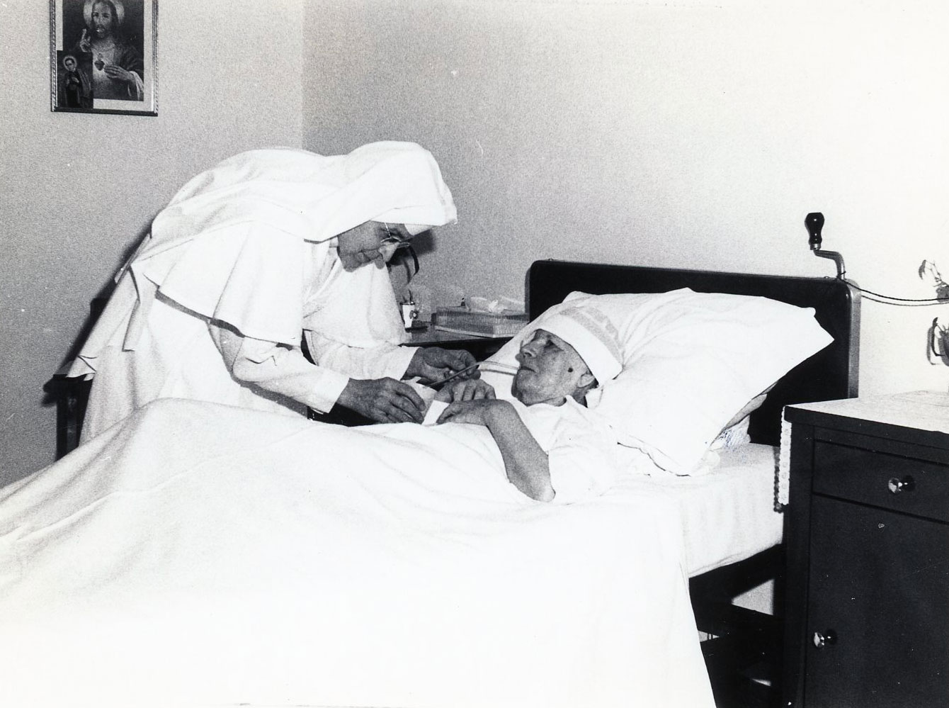 Sister Mary Leon Bichsel, wearing a white modified habit, cares for Sister Adolpha Zwick, who is lying in bed, ca 1963-1969.