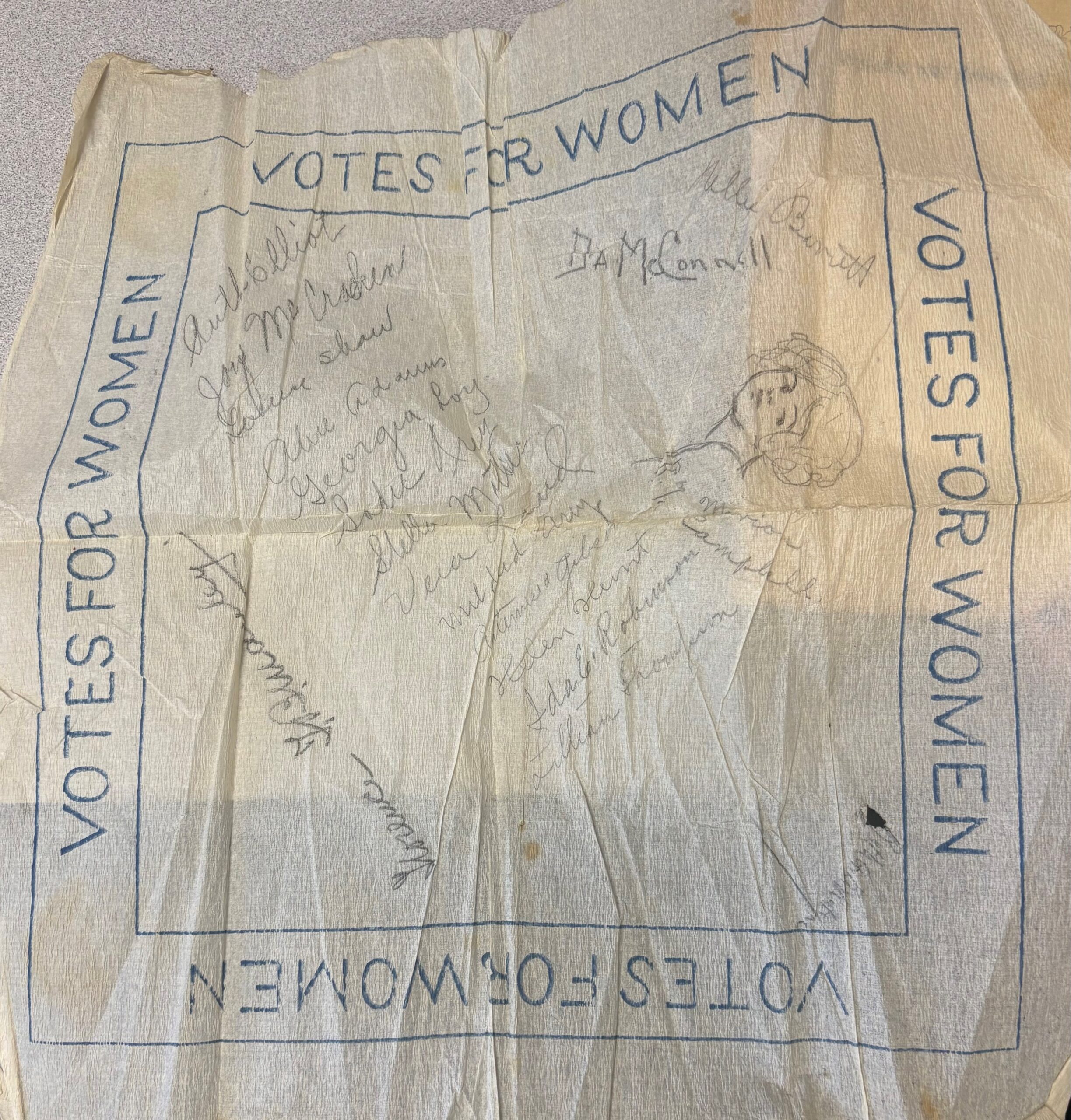 A piece of tissue paper that reads, “Votes for Women” and contains a drawing of a young woman and the signatures of a number of women.